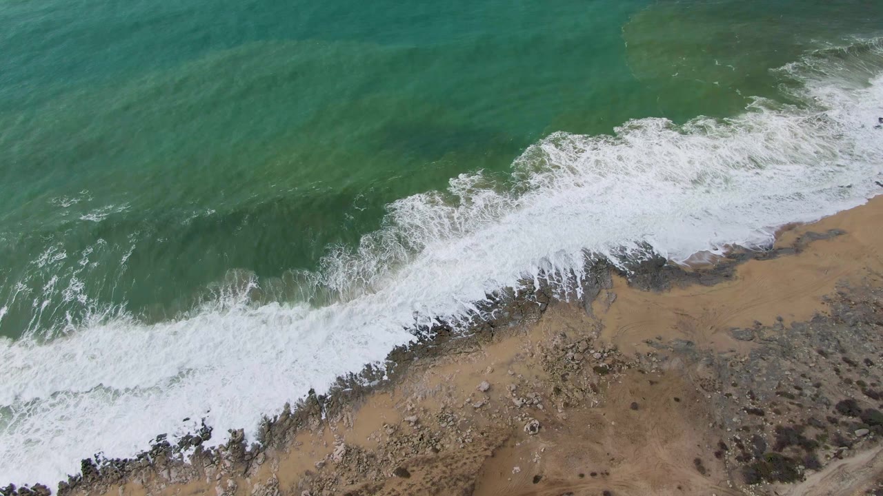 Overhead view at Punta Gorda, Baja California Sur, where green ocean water breaks into white foam over a tan sandy and rocky shoreline, with no visible people.