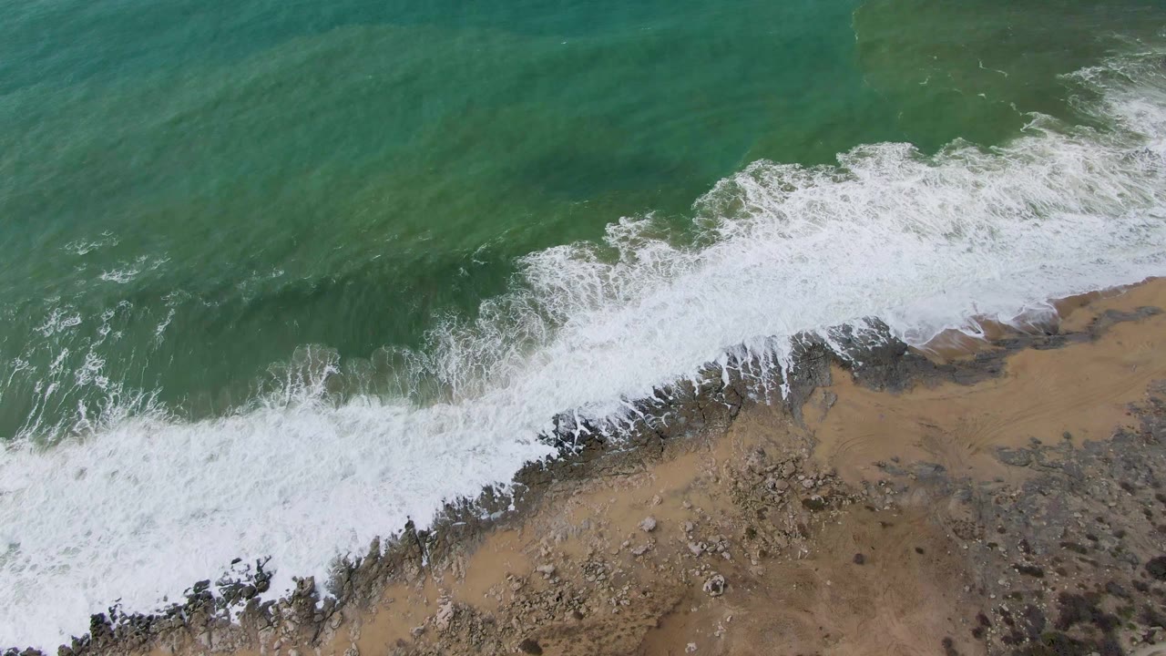 At one second, white surf thickens along the Punta Gorda rocky shoreline while green water remains above the diagonal break, with no visible people.