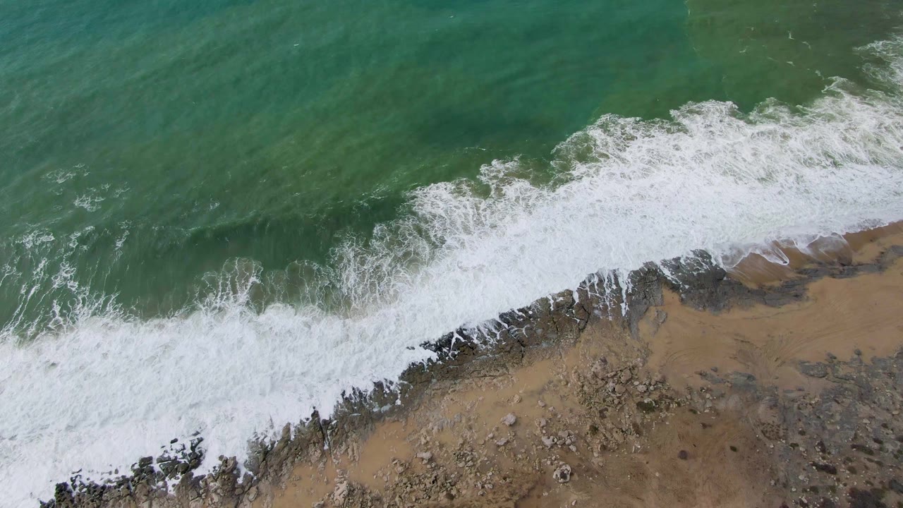 At two seconds, the aerial view shows a wider sheet of white foam crossing the sandy and rocky Punta Gorda coast under green ocean water, with no visible people.