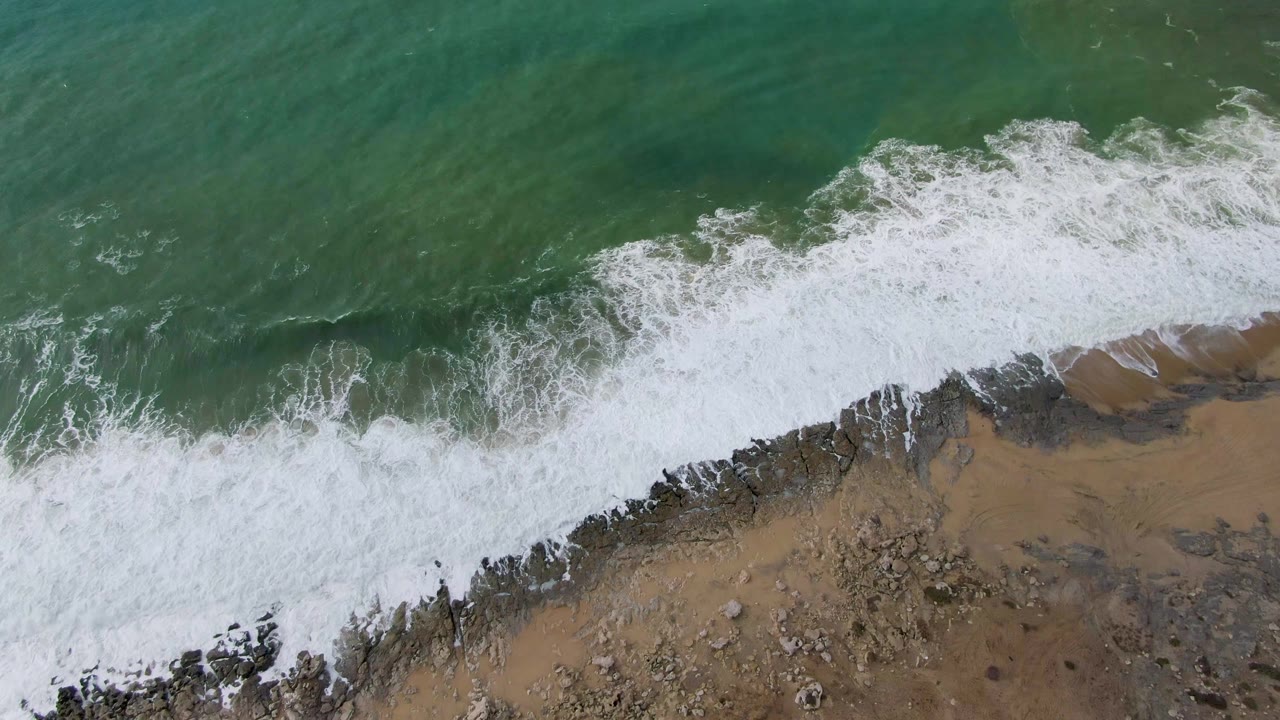 Foam washes diagonally along a rocky tan shoreline from an overhead angle.