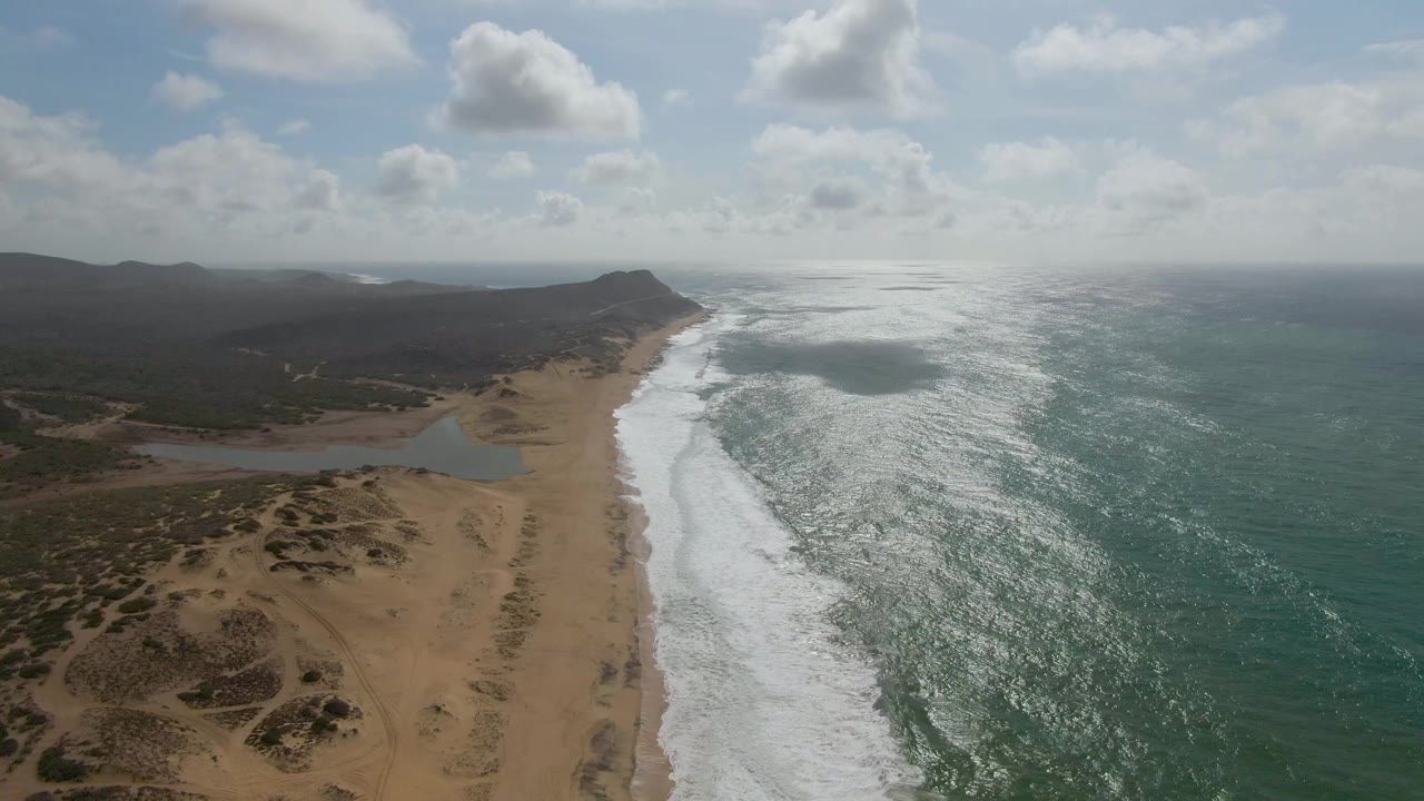 Aerial view at 0:01 over Punta Gorda, Baja California Sur, with foamy wave bands advancing along the sandy shoreline, the lagoon set back from the beach, dark low hills behind it, sunlit ocean texture, and no visible people.