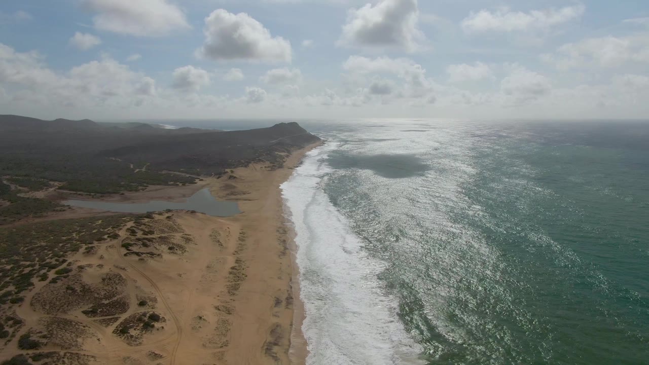 Mid-clip aerial frame from Punta Gorda, Baja California Sur, showing bright reflected light on the ocean, white surf curling onto the sand, lagoon water left of the beach, low inland hills, scattered clouds, and no visible people.