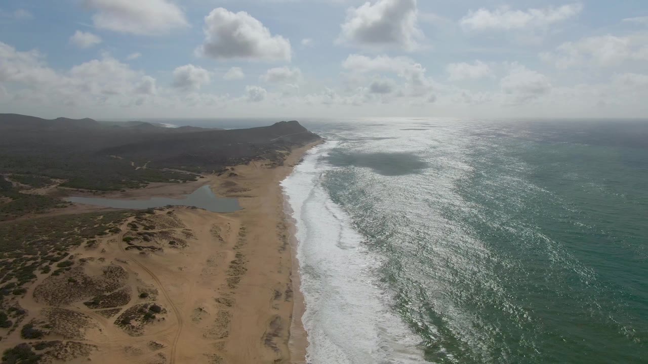 At 0:03, the aerial view over Punta Gorda, Baja California Sur, holds the shoreline diagonally through frame, with dunes in the foreground, a lagoon near the beach, continuous white surf, open ocean, hazy low hills, and no visible people.