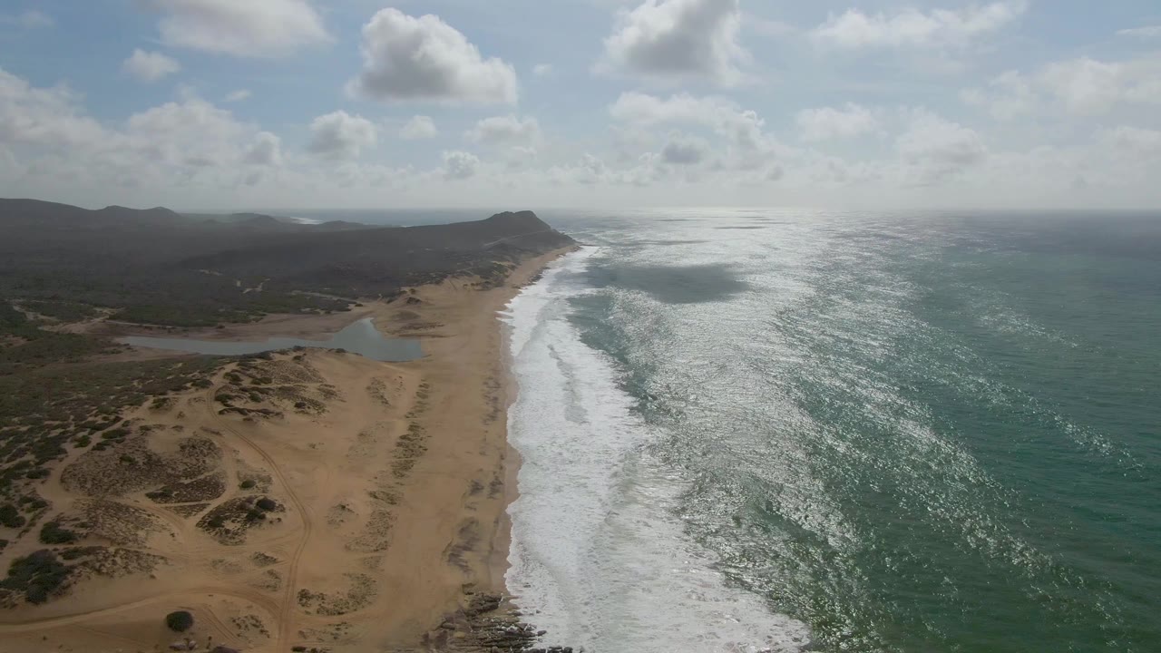 Aerial frame over Punta Gorda, East Cape, showing white surf running beside sandy dunes, green-blue ocean, distant dark hills, partly cloudy sky, and no visible people.