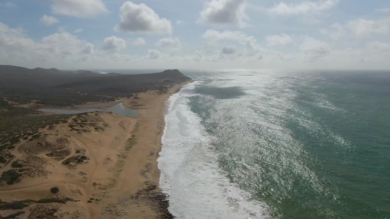 Punta Gorda, East Cape aerial frame with bright reflected light on the ocean, a long band of whitewater along the sand, low dunes in the foreground, hazy hills, and no visible people.