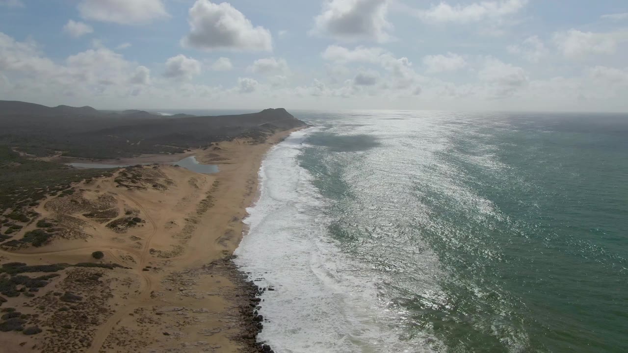 Aerial frame from Punta Gordo, Baja California Sur, showing white surf washing along a sandy beach beside dunes and distant dark hills, with no visible people.