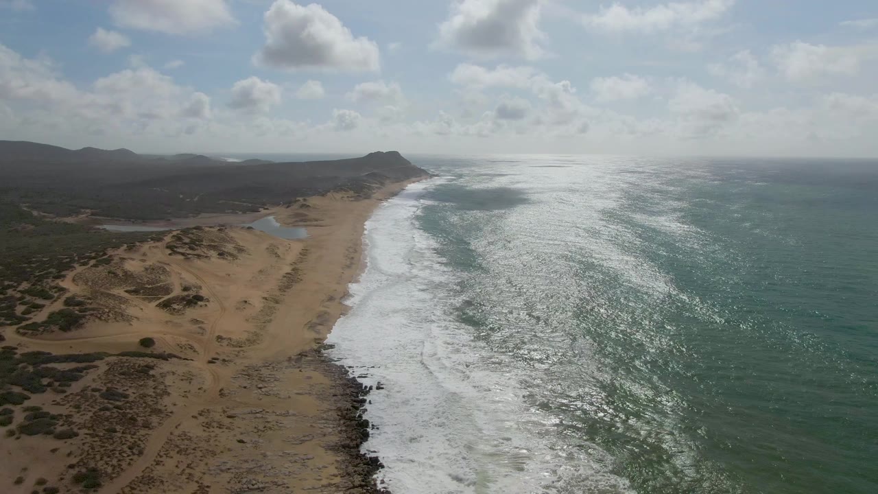 Diagonal coastline with white waves, sandy dunes, and a partly cloudy sky.