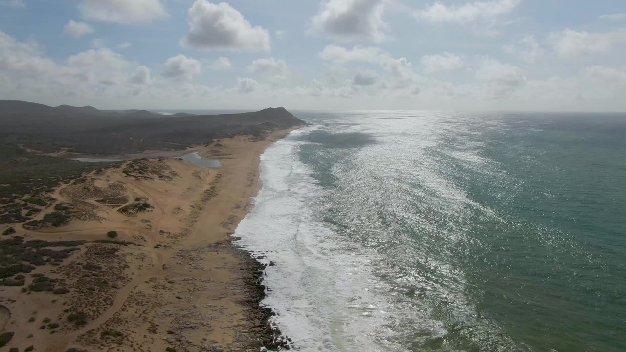 Aerial frame from Punta Gordo showing sunlit green-blue water, a long white foam band, sandy dunes, inland scrub, distant hills, and no visible people.
