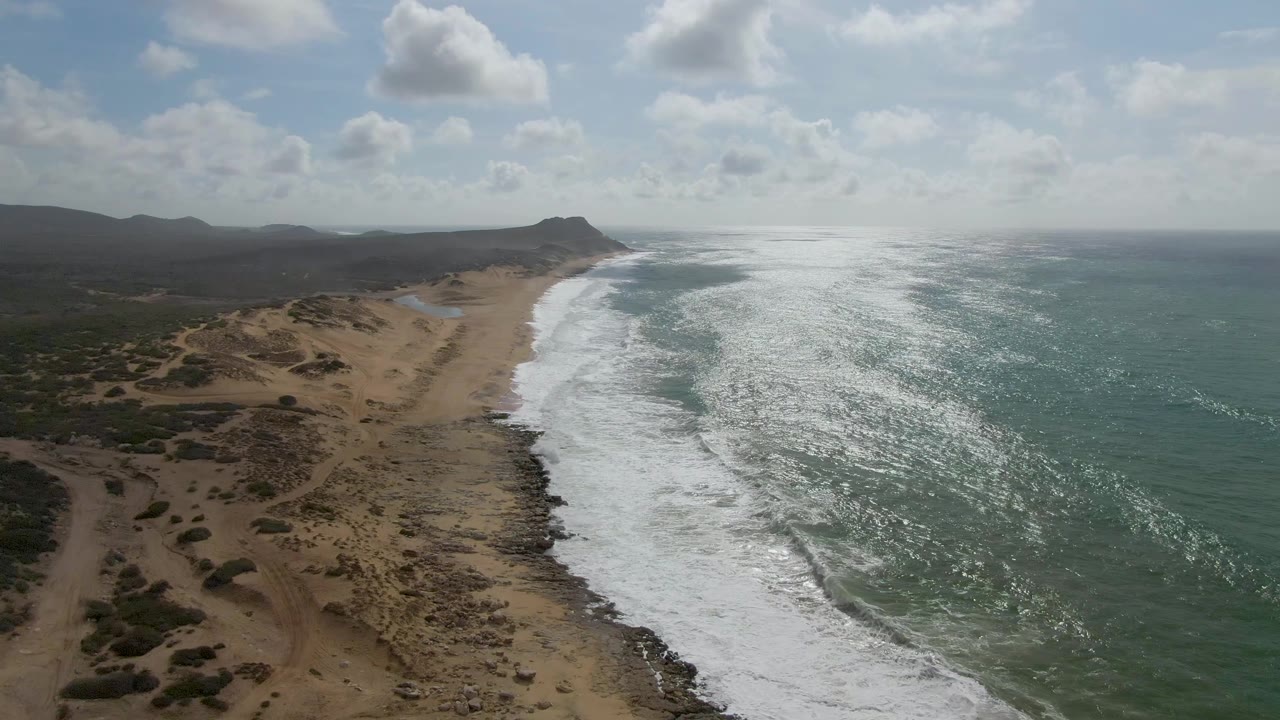 Aerial view at Punta Gorda showing a brighter band of white foam pushing up the shoreline, tan dune ridges and scrub on the left, green-blue water on the right, hazy hills in back, and no visible people.