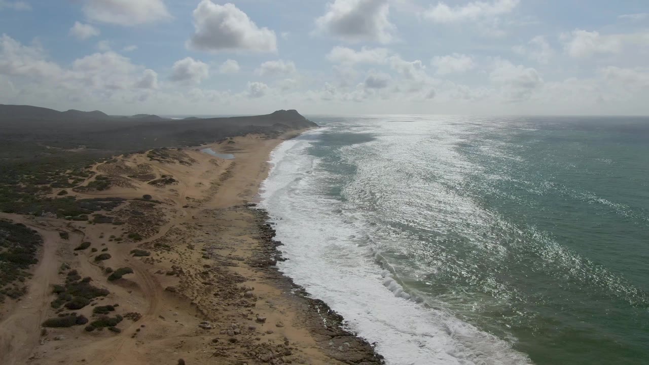 Aerial frame from Punta Gorda with sun glare across the ocean surface, a broad white surf band along the shore, tan sand and low scrub to the left, dark hills on the horizon, scattered clouds, and no visible people.