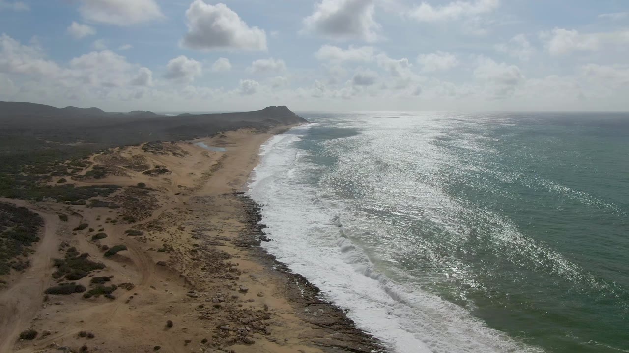 Punta Gorda aerial frame with the beach curving through the center, waves spreading white foam over the shore, scrub-covered dunes in the lower left, open green water to the right, distant hills, clouds, and no visible people.