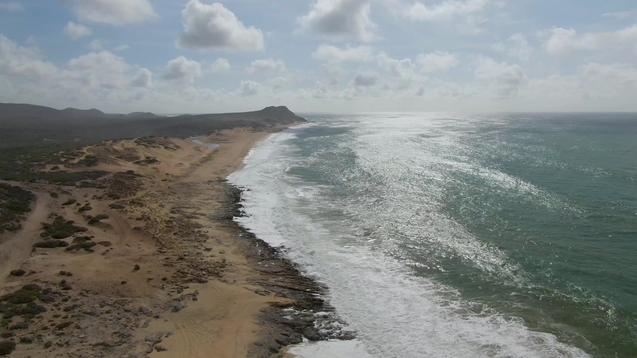 Aerial view of Punta Gorda East Cape coastline with white surf curling along a sandy and rocky beach, scrub-covered land to the left, green-blue ocean to the right, and no visible people.
