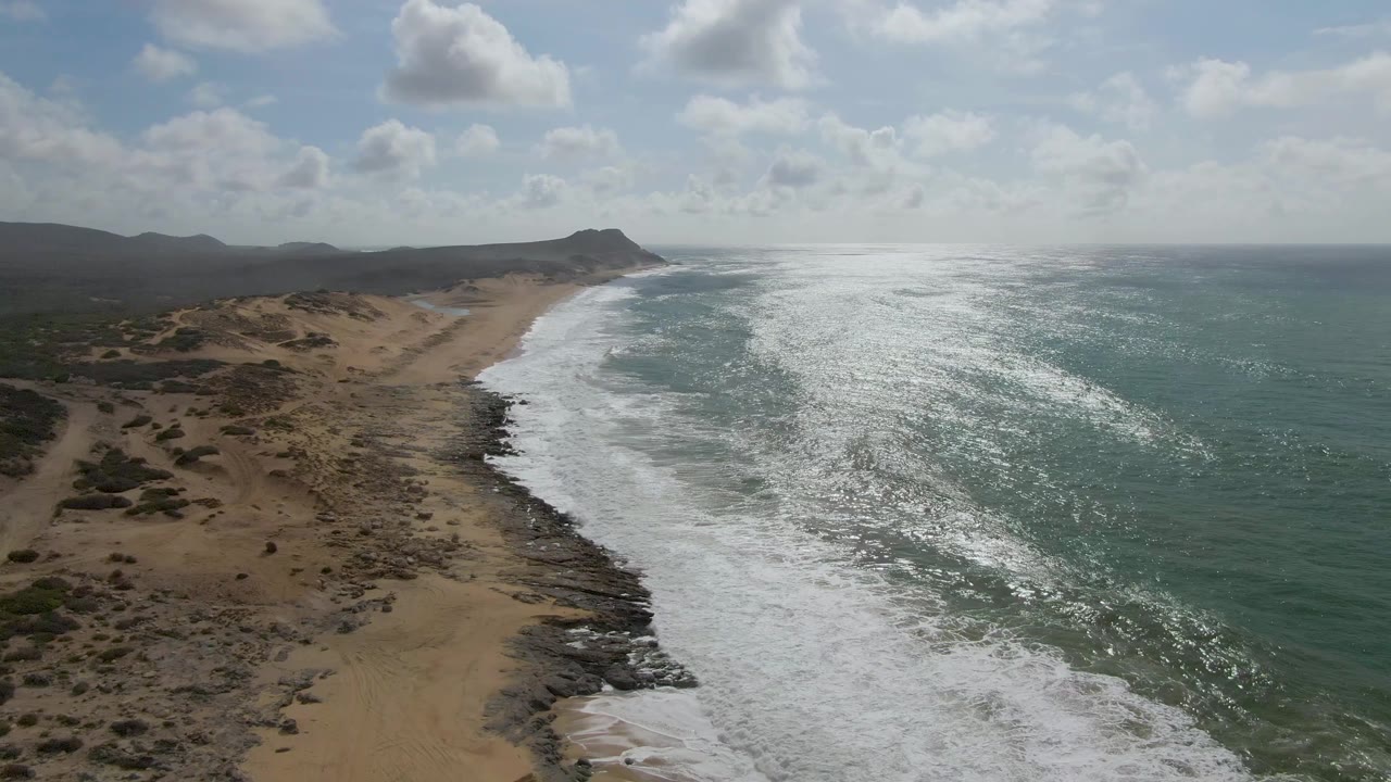 Drone-style aerial frame of Punta Gorda East Cape with a broad white foam band reaching farther down the sandy shore, rocky patches near the waterline, distant hills, and no visible people.