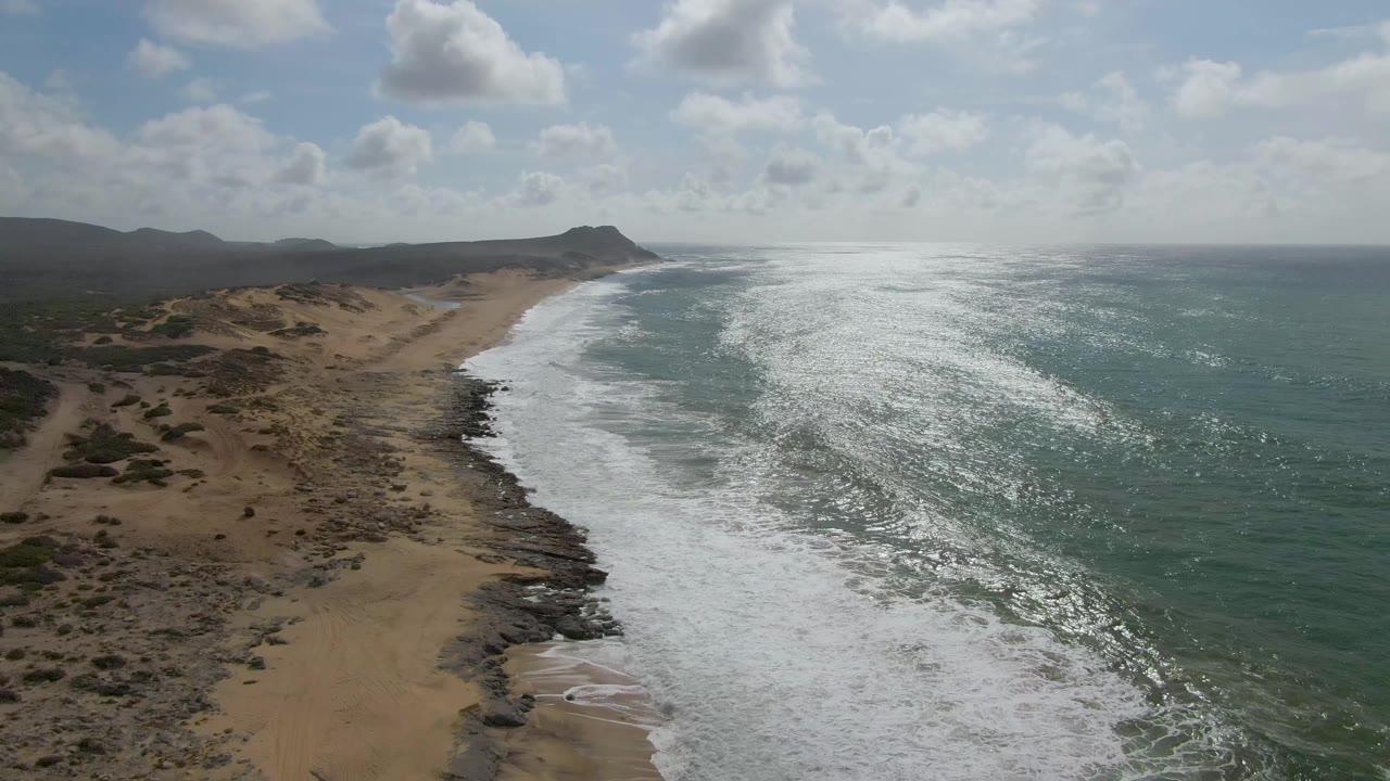 Aerial coastal view in Punta Gorda East Cape showing strong sunlight glitter on the ocean, a curving white surf line, tan beach, scattered rocks, scrubland, and no visible people.