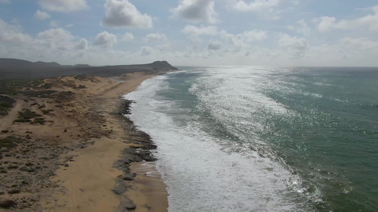 Wide aerial beach view with surf, rocks, and low hills