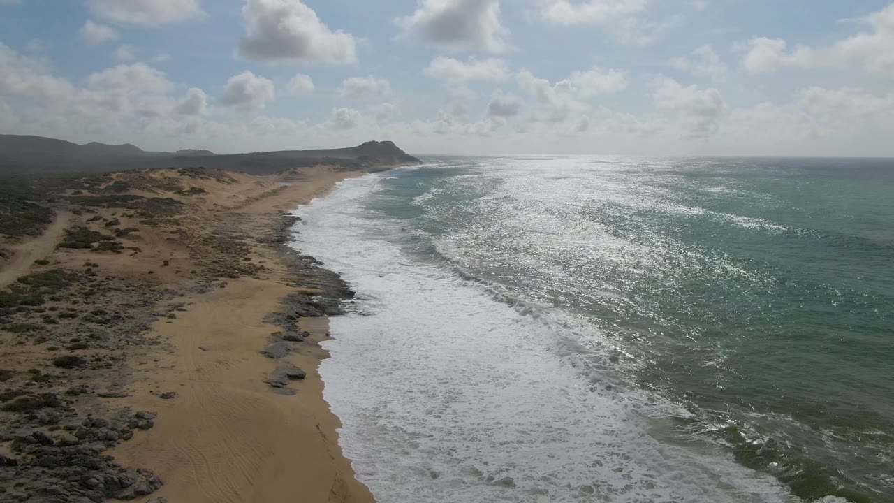 Aerial frame over Punta Gorda East Cape, Baja California Sur, showing white surf running along a sandy beach with rocky outcrops, distant hills, cloud cover, and no visible people.