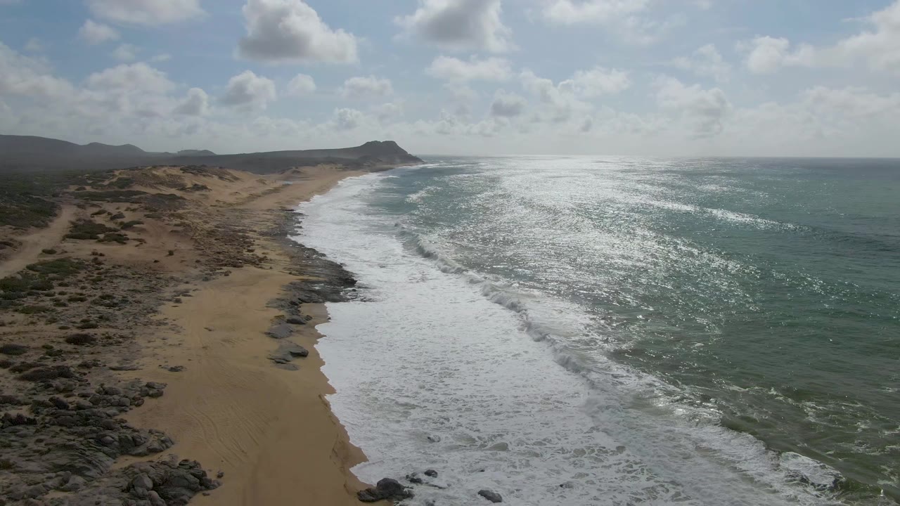 White waves wash along a curved sandy beach with rocks inland.