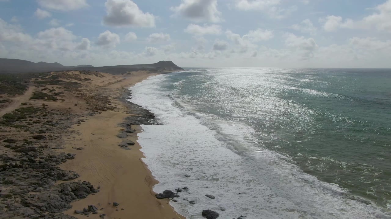 Elevated frame over the Punta Gorda East Cape coastline, with breaking surf filling the right side, sand and rock formations along the left, distant landforms, and no visible people.