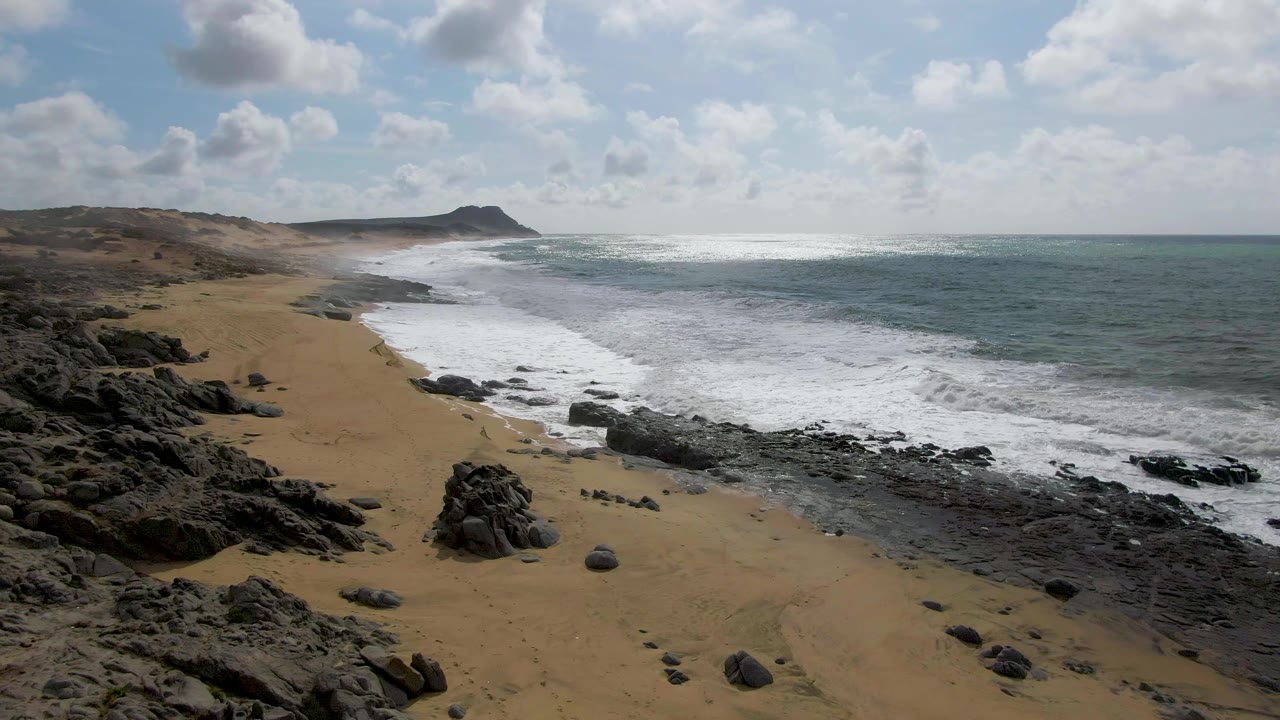 At 0:01 in Punta Gorda, Baja California Sur, white foam spreads farther across the shoreline near dark rock outcrops, with empty sand and no visible people.