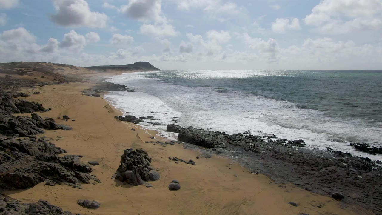 At 0:02 in Punta Gorda, Baja California Sur, the surf line curves along the beach while dark rocks sit in the foreground and a low headland remains visible in the distance, with no visible people.