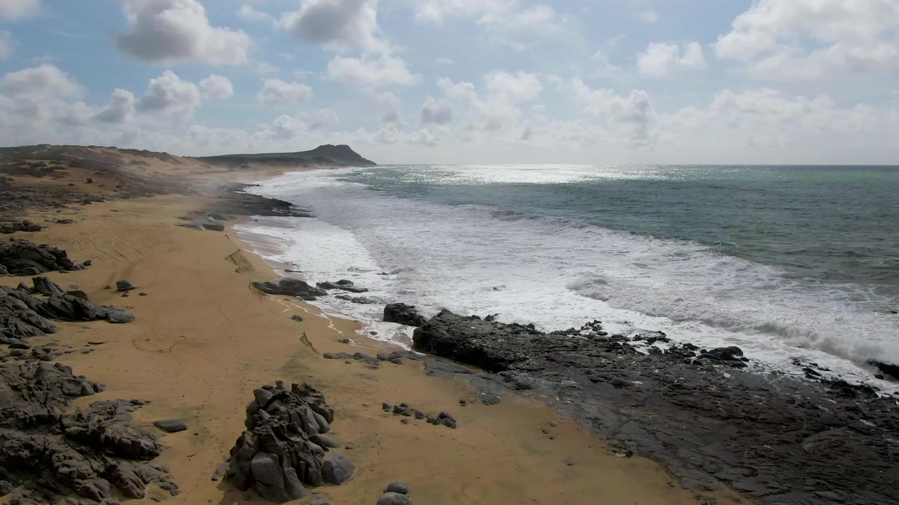At 0:03 in Punta Gorda, Baja California Sur, bright white waves wash across the rocky edge of a broad sand beach under scattered clouds, with no visible people.