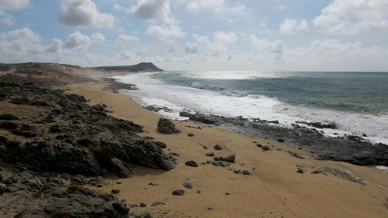 Rocky sandy shoreline with waves breaking under scattered clouds