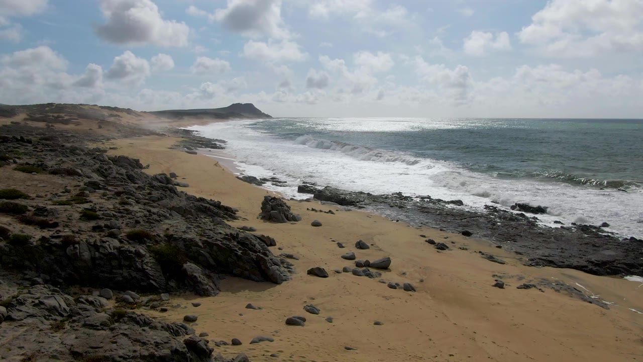 Punta Gorda East Cape coast with foam advancing across the midground, dark rocks in the foreground, open ocean to the right, and no visible people.