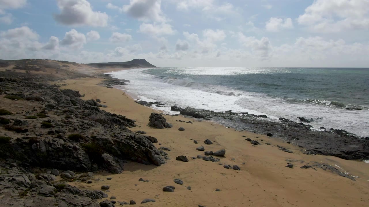Elevated Punta Gorda East Cape view with a broad band of whitewater near shore, dark foreground rocks, tan beach, low headland on the horizon, and no visible people.