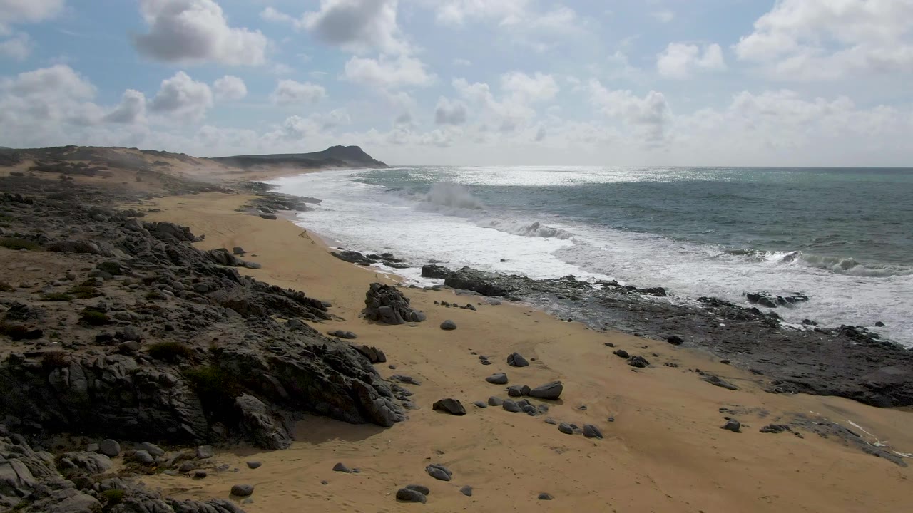 Punta Gorda East Cape shoreline with darker foreground rocks, sand curving toward the headland, churning surf near shore, scattered clouds above, and no visible people.