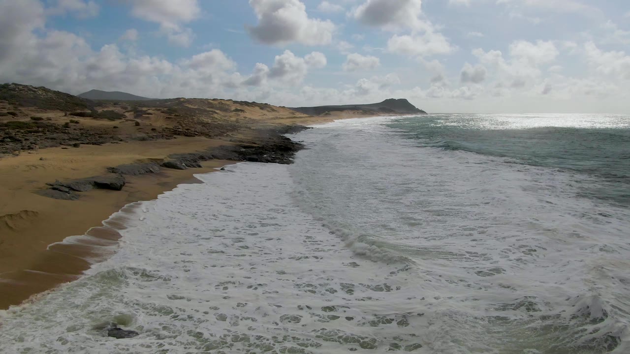 At 0:00, foamy surf spreads across the foreground at Punta Gorda, Baja California Sur, with tan beach, dark rocks, low hills, scattered clouds, and no visible people.