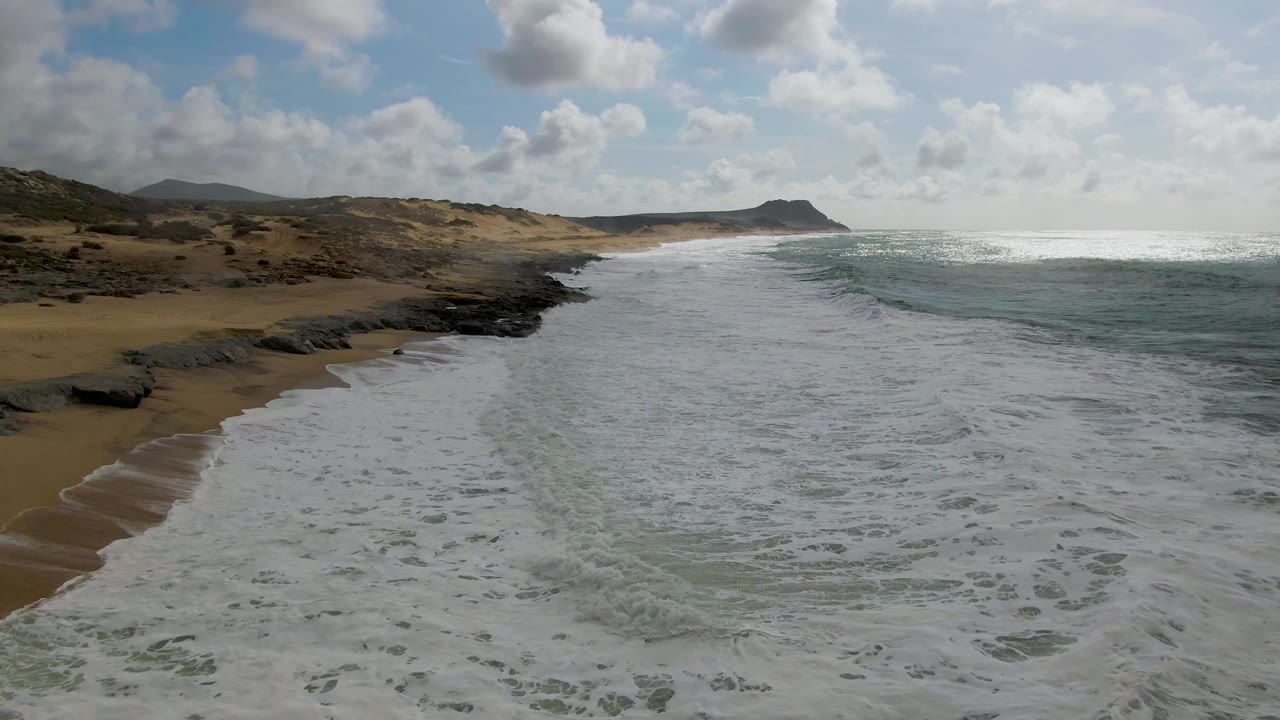 At 0:01, the surf line has advanced along the left beach at Punta Gorda, with blue-green water to the right, rocky shoreline ahead, clouds above, and no visible people.