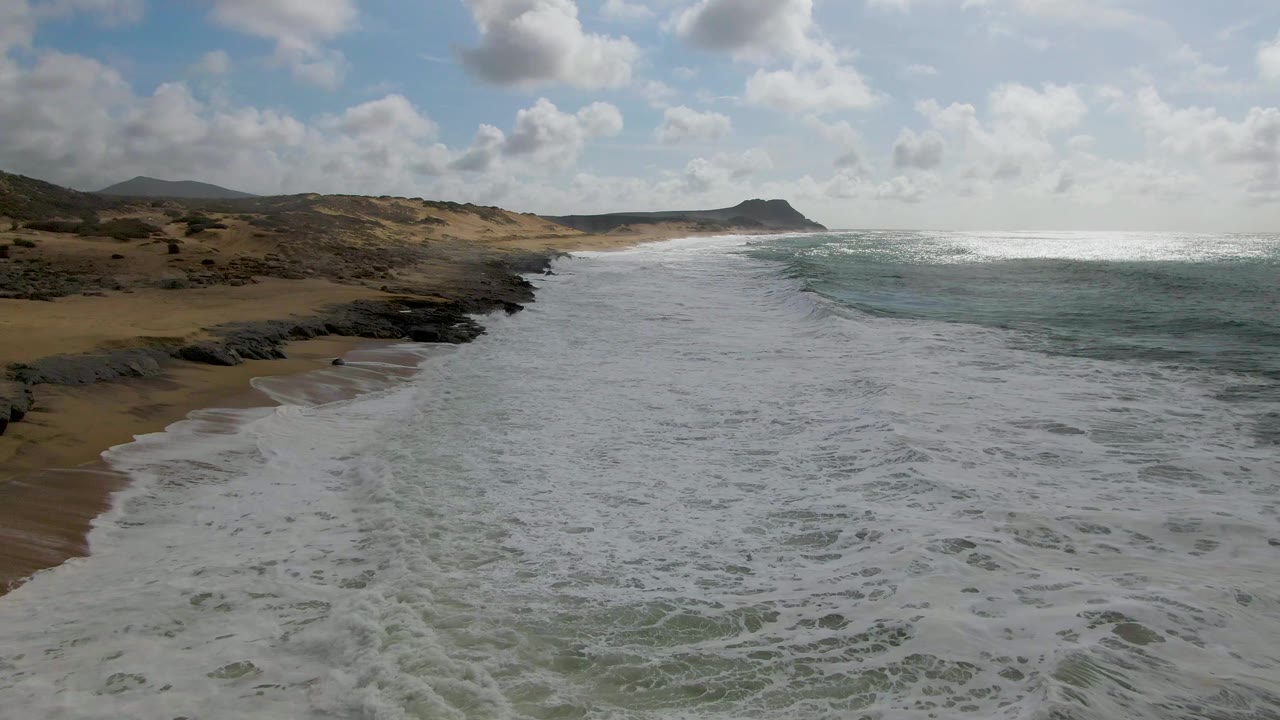 At 0:02, broad white foam fills more of the lower frame at Punta Gorda, while the sandy left edge, dark rock ledges, distant headland, and no visible people remain in view.