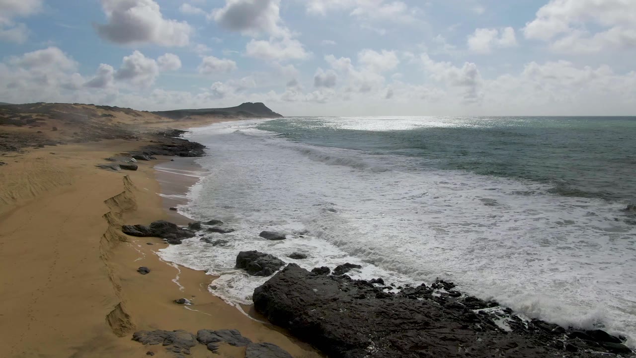 At 0:00 in Punta Gorda, Baja California Sur, white surf spreads toward dark foreground rocks along a sandy beach, with a low headland in the distance and no visible people.