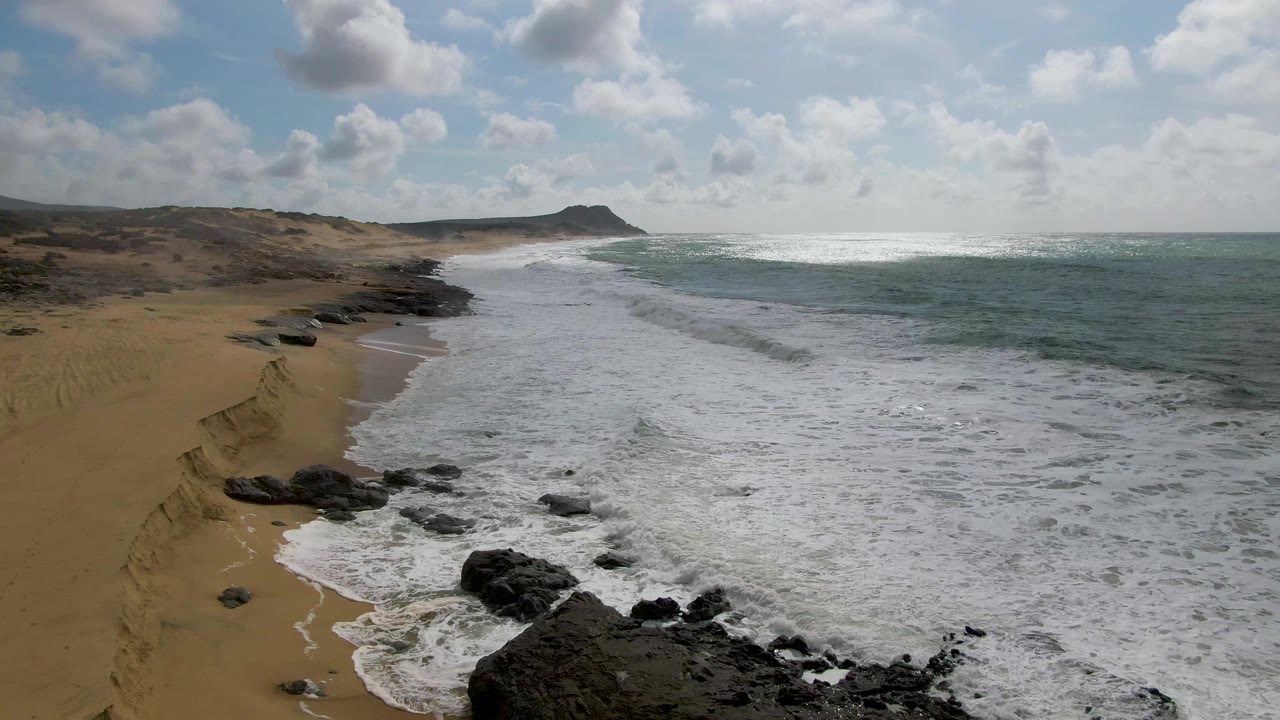 At 0:01 in Punta Gorda, Baja California Sur, the foamy wash reaches higher along the sand beside dark rock outcrops, while the ocean and clouded sky fill the right side with no visible people.