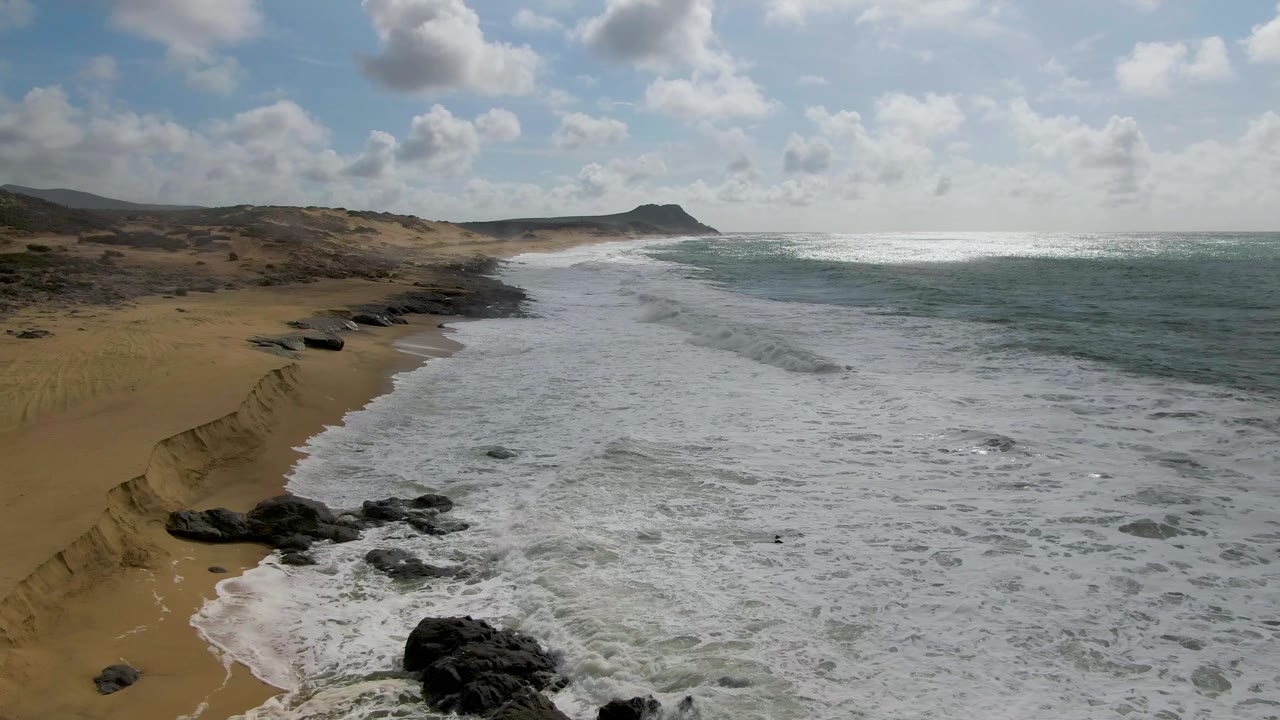 At 0:02 in Punta Gorda, Baja California Sur, broad white surf covers much of the beach edge, dark rocks sit near the lower frame, and a low headland remains visible beyond the coastline with no visible people.
