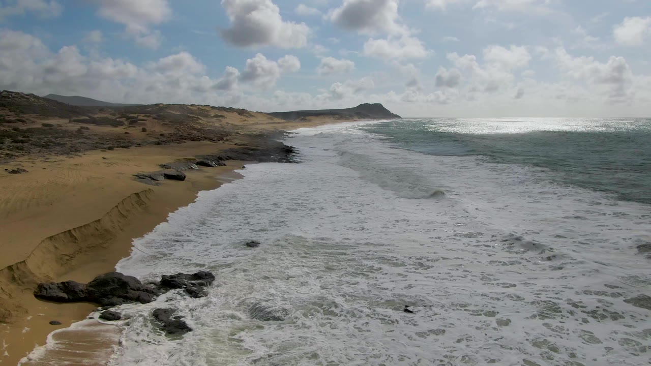 At 0:03 in Punta Gorda, Baja California Sur, a sandy cut bank and beach run along the left side while whitewater pushes inward from the ocean, with rocks at the lower edge and no visible people.