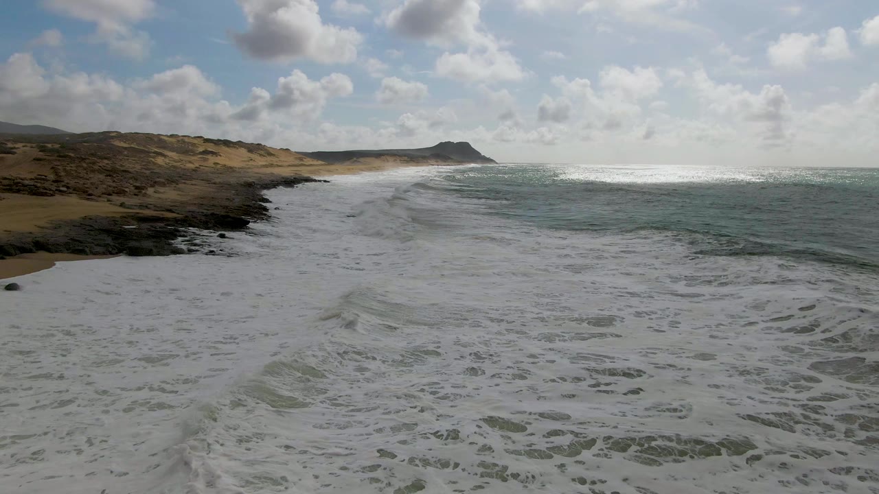 Foamy surf spreads across a sandy Punta Gorda East Cape shoreline, with low dunes and dark rocks at left, open ocean at right, partly cloudy sky, and no visible people.