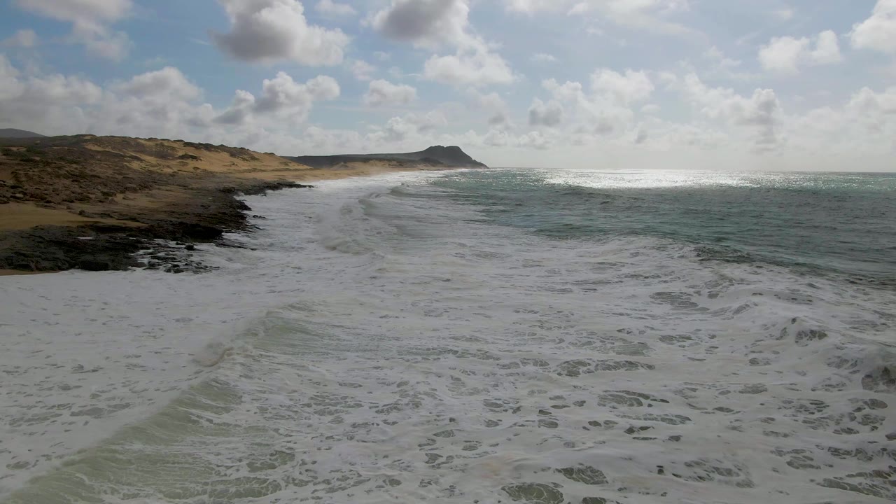 A fresh line of white surf pushes shoreward at Punta Gorda East Cape, with dark rocky edges on the left, sandy dunes beyond, blue-green water to the right, and no visible people.