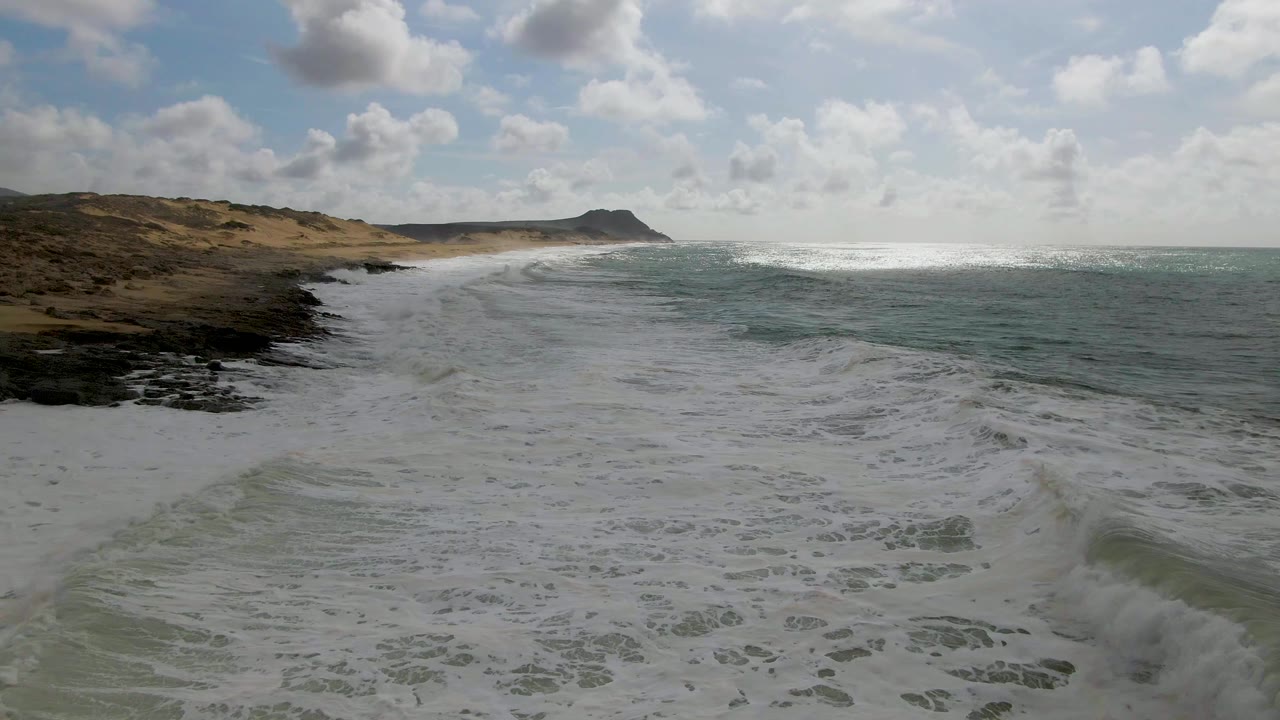 White foam bands cover the foreground water along the Punta Gorda East Cape beach, while dunes and rocks sit left of frame, open sea fills the right side, and no visible people appear.