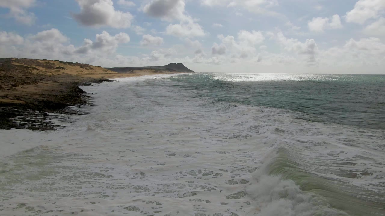At 0:03, the Punta Gorda East Cape surf flattens into broad whitewater across the foreground, with a sandy beach and low dunes on the left and blue-green ocean on the right, no visible people.