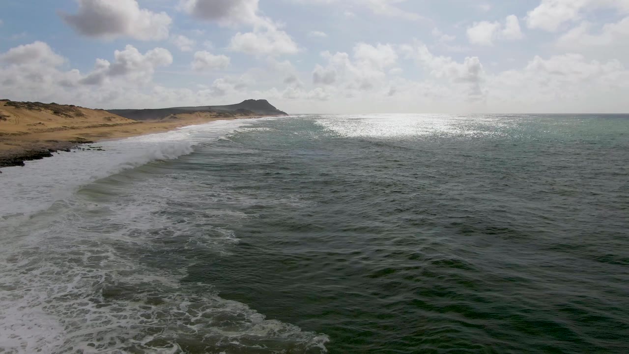 Opening aerial frame of Punta Gorda East Cape, Mexico, with white surf washing diagonally along the sandy shoreline, green-blue water in the foreground, a distant headland, and no visible people.