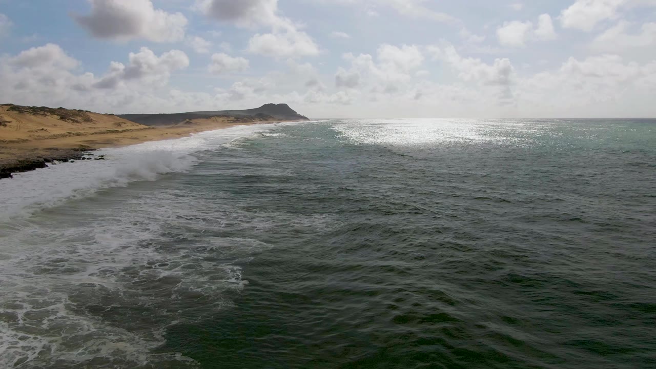 White foam spreads from a breaking wave beside a sandy beach and low dunes.