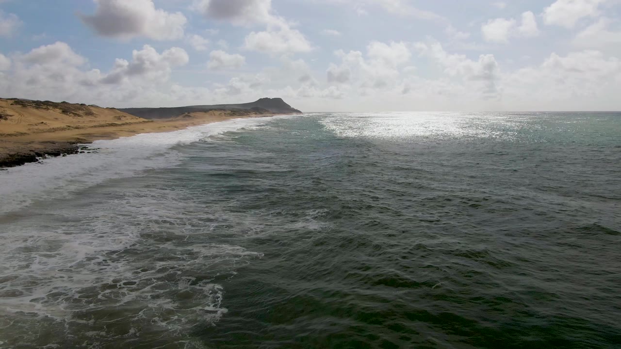At 0:02, the Punta Gorda East Cape shoreline remains on the left as white foam trails behind a breaking wave, open water fills the right side, and no visible people appear on the beach or sea.
