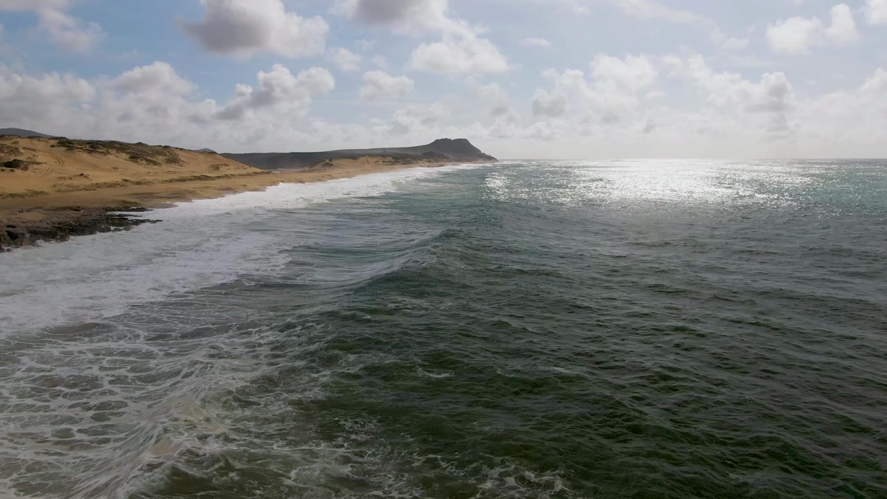 Opening aerial view at Punta Gorda, East Cape, Baja California Sur, with green-blue water, white surf along the sandy beach, low dunes, distant headland, and no visible people.