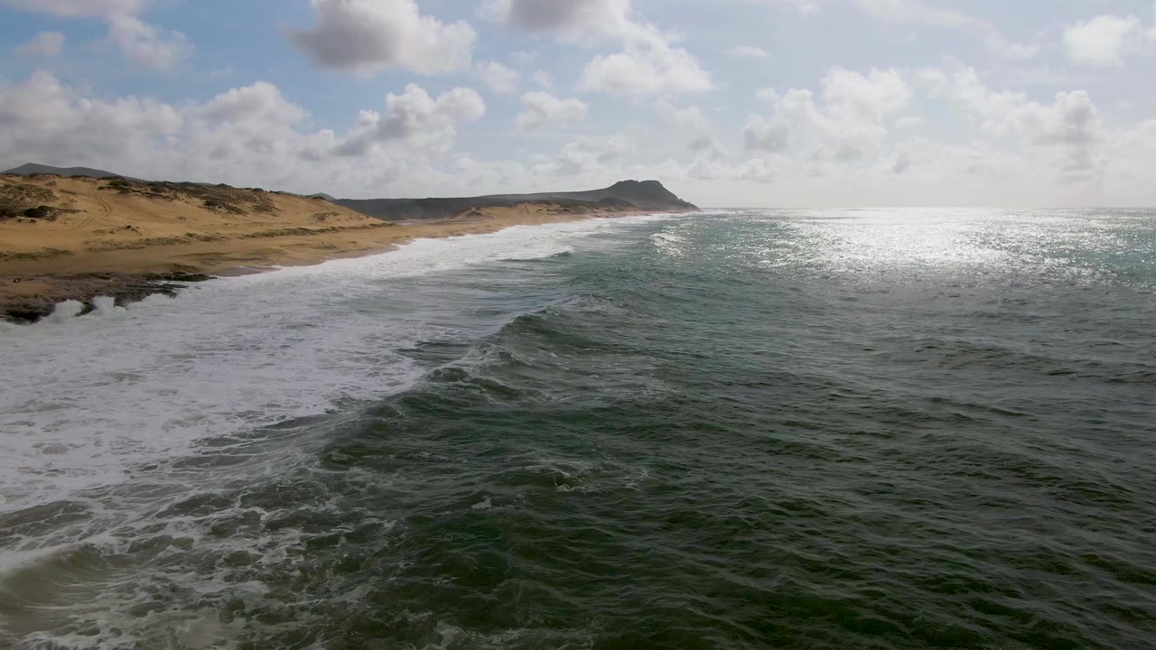 At 0:02, a diagonal wave crest advances toward the Punta Gorda beach, with wind-textured green water in front, tan dunes behind, and no visible people.