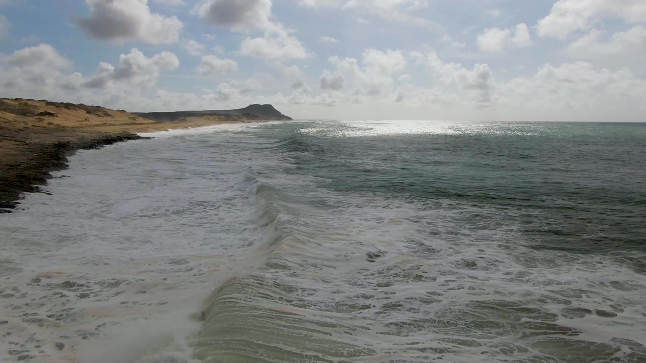 At Punta Gorda, Baja California Sur, white surf spreads across wet sand below a partly cloudy sky, with green-blue ocean to the right, a distant headland ahead, and no visible people.