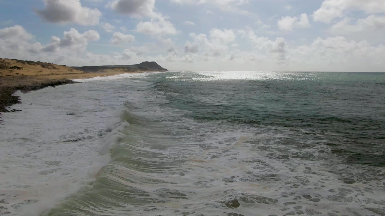 At Punta Gorda, Baja California Sur, the wave edge reaches farther up the sandy beach while the bright ocean surface and low headland remain in the distance, with no visible people.