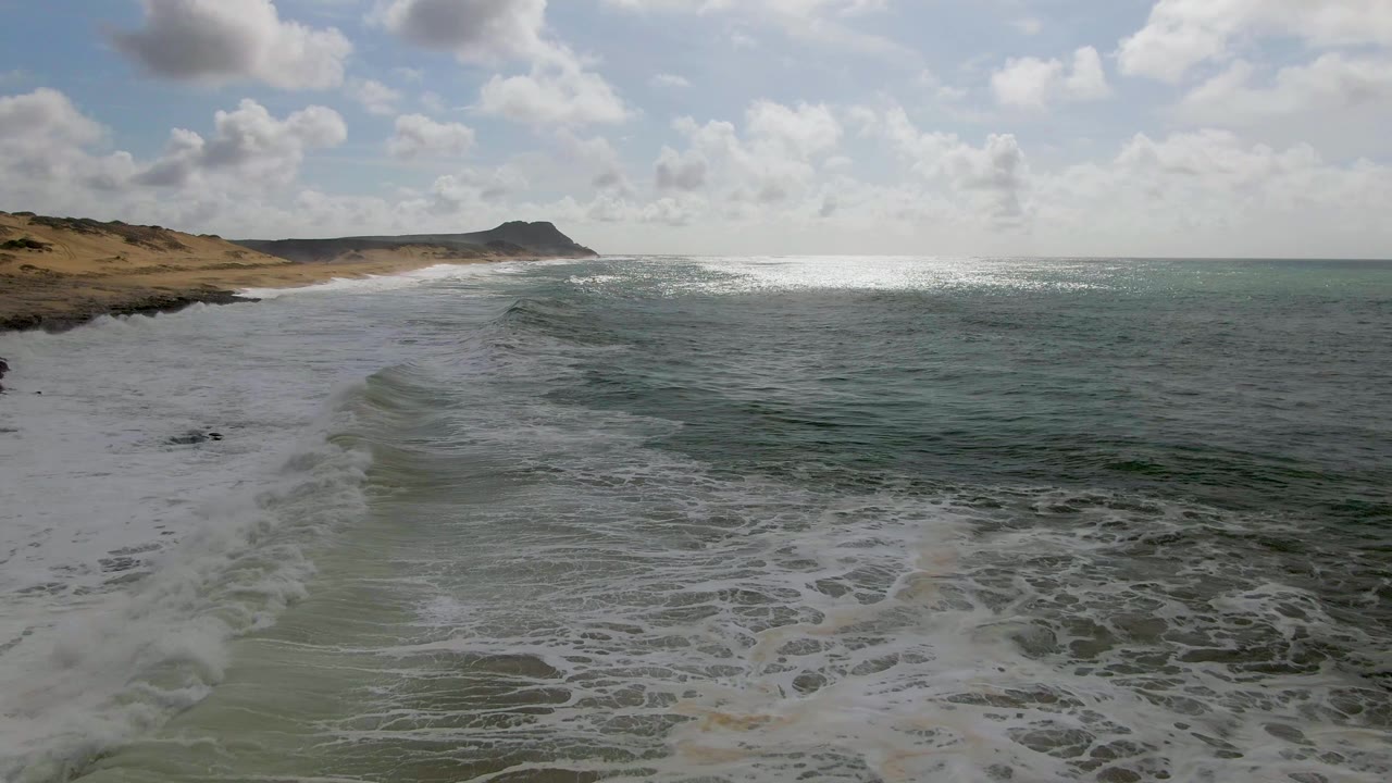 At Punta Gorda, Baja California Sur, foamy water covers more of the foreground shore, the ocean stays dark green to the right, and the headland sits under broken clouds with no visible people.