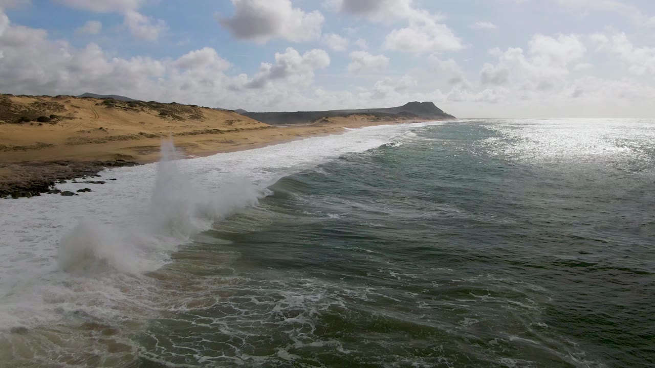 At Punta Gorda on the East Cape of Baja California Sur, a wave throws white spray near dark shoreline rocks while green-blue water fills the foreground and no people are visible.