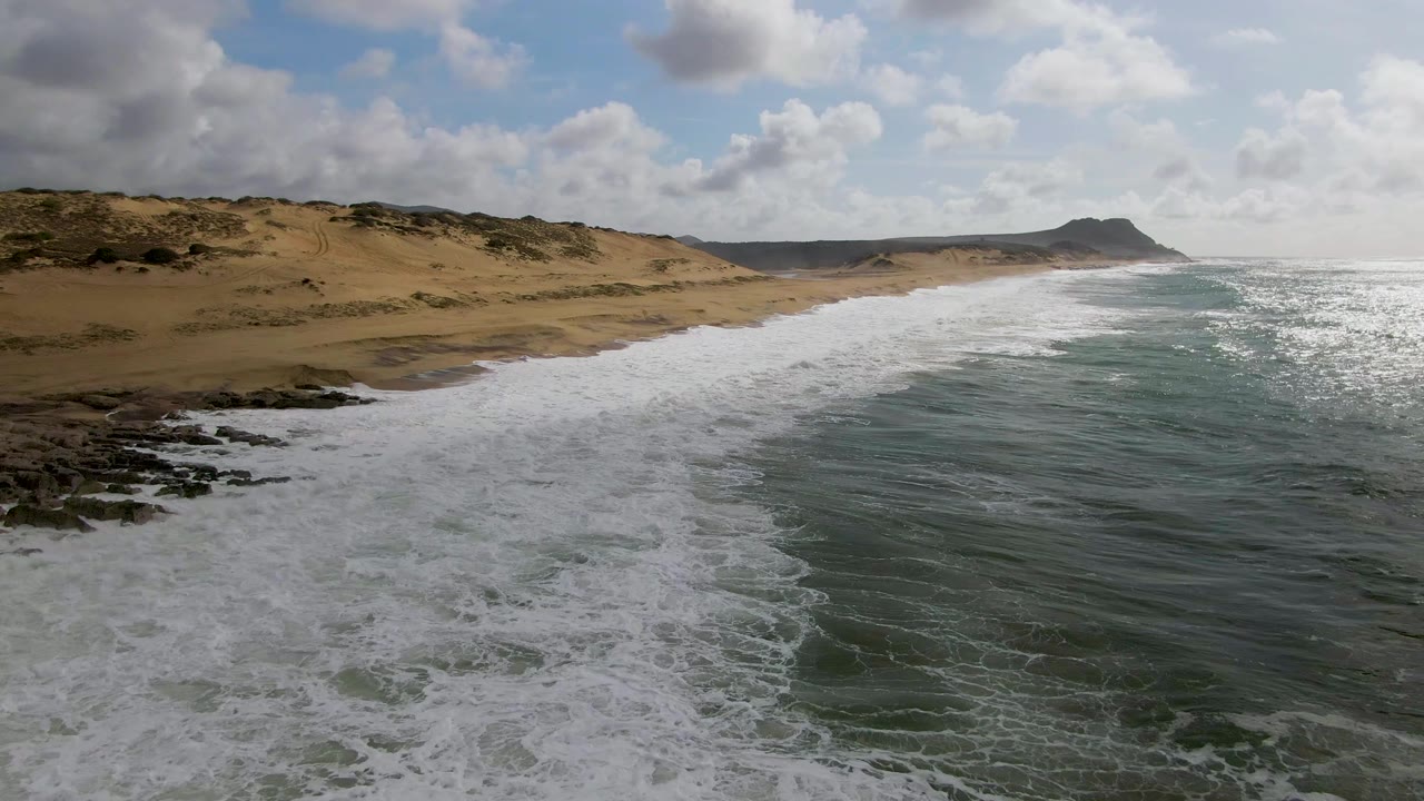 Aerial view at Punta Gorda, Baja California Sur, with white surf spreading over blue-green water toward a sandy beach, rocky foreground, low dunes, a distant headland, and no visible people.